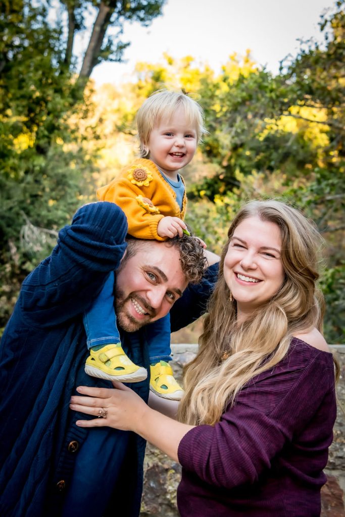 outdoor family portrait of parents with toddler on shoulders by photographer Shelly Rivoli