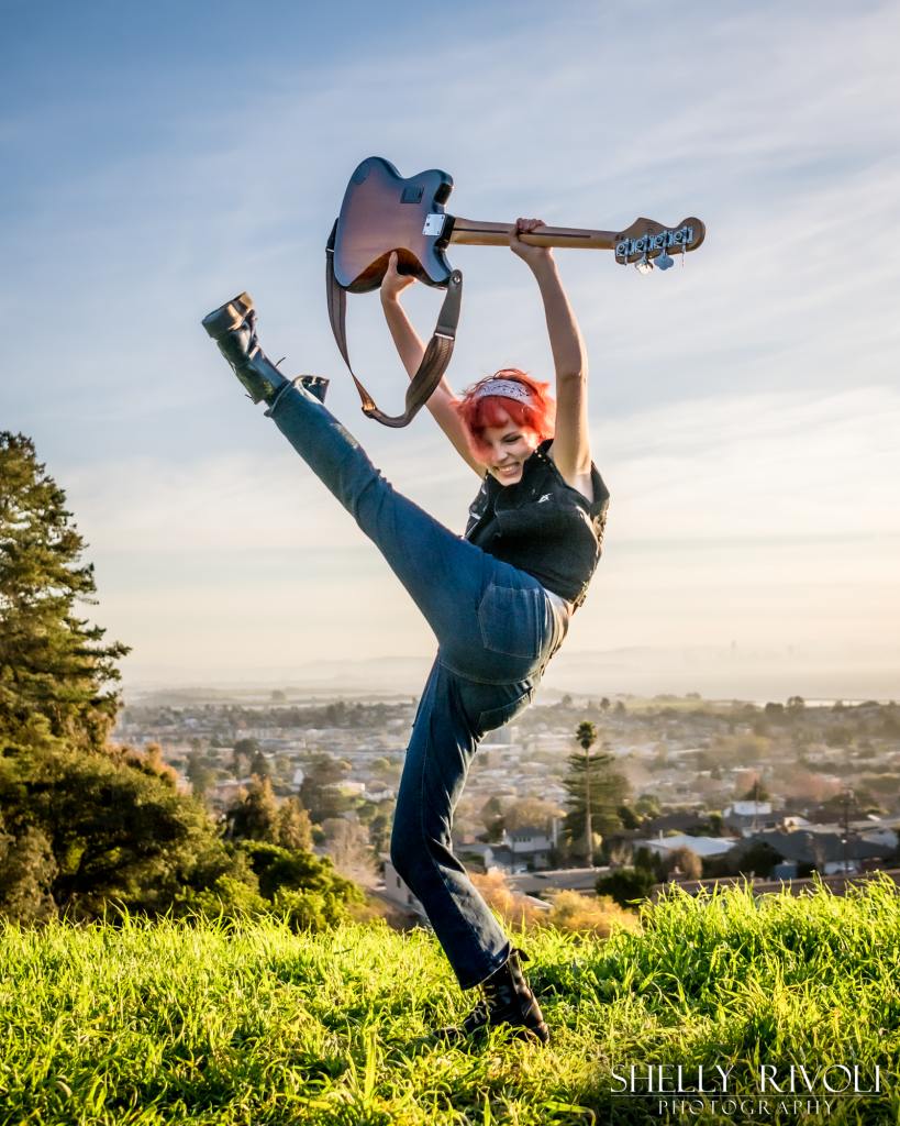 Senior portrait of punk teen girl kicking leg high with bass guitar in air by photographer Shelly Rivoli